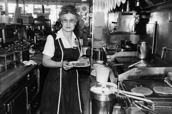Esther, a Rennebohm Drug Store restaurant employee, holds an order of eggs and toast while keeping watch on potatoes and pancakes cooking on the grill. She is wearing a "Colossal Cupcake Fever" button.