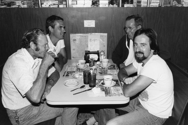 Four men in informal attire on their coffee break in an unidentified Rennebohm Drug Store restaurant. They are sitting in a booth identified as a no smoking area. Their booth directly adjoins the drug store and, ironically, cartons of cigarettes for sale can be seen at the top of the photograph.