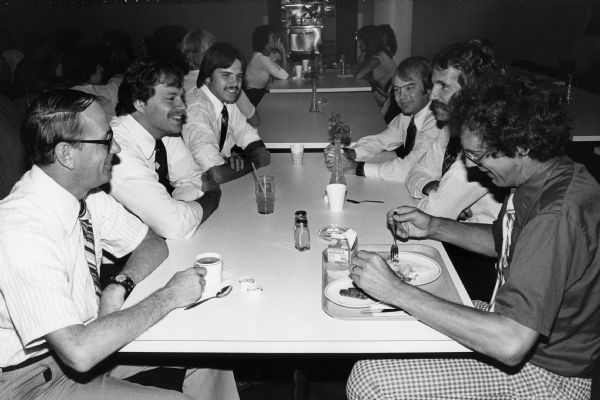 A group of men, all in shirt sleeves and ties, sit at a table at the Rennebohm Drug Store No. 6 restaruant in the First Wisconsin Bank on Capitol Square. All are enjoying a beverage, while the man on the right is eating eggs and sausage.