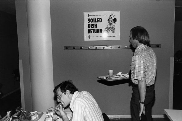 A sign reading "Soiled Dish Return" directs a man bussing his own table at the Rennebohm Drug Store No. 6 restaurant in the First Wisconsin Bank on Capitol Square. Another man can be seen eating at a table.