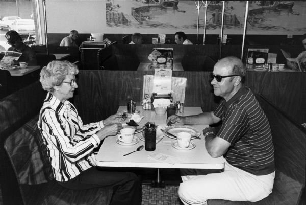 A couple eats breakfast at an unidentified Rennebohm Drug Store restaurant. Other diners can be seen in the seating area behind them.