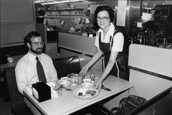 A waitress serves ham and eggs to Steven O. Kimbrough at an unidentified Rennebohm Drug Store restaurant. Chuck Patch, Kimbrough's breakfast companion, has gotten up to take the photograph, leaving his camera bag on the seat. Patch and Kimbrough were in the midst of a personal project to eat breakfast and photograph all of the Rennebohm restaurants. An unusual feature at this restaurant were booths with pass-through access from the waitresses' work area.
