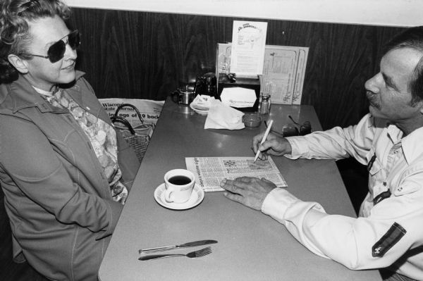 Two people take a break at an unidentified Rennebohm Drug Store restaurant: the woman enjoys a cup of coffee, her companion works on a crossword puzzle.
