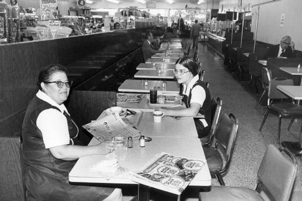 Two Rennebohm employees take a break to read the newspaper. Although unidentified, this photograph by Chuck Patch, is thought to be the Rennebohm Drug Store No. 7, 901 University Avenue. This restaurant featured both cafeteria and counter service.