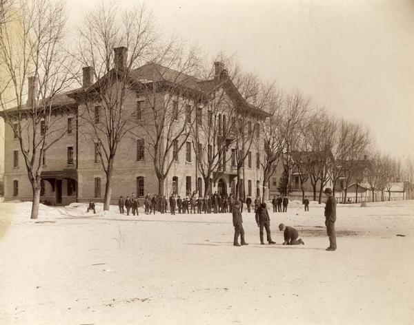 Students gather in the schoolyard at the Wisconsin Industrial School for Boys. A group of four boys is seen in the foreground, while dozens of others stand outside a school building in the background.