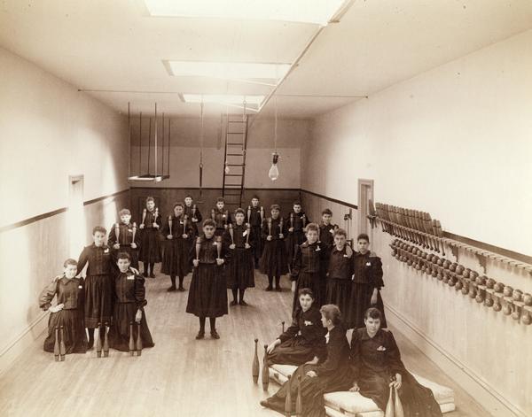 Elevated view of group of female students posing with Indian clubs in a gymnasium at the State School for the Deaf.