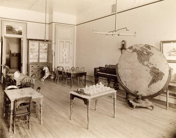 Interior of the State School for the Blind, perhaps a study or room for recreation. Included in the room are several tables with small models, a piano, and a large map of the Americas with geographic representations indicated in relief.