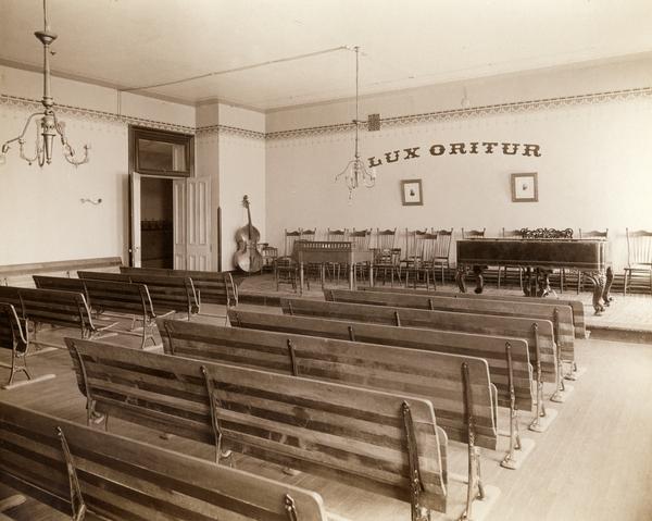 Music recital room at the State School for the Blind with seven rows of seating, a piano and cello. Adorning the front wall is the Latin phrase "Lux Oritur" ("A light arises").