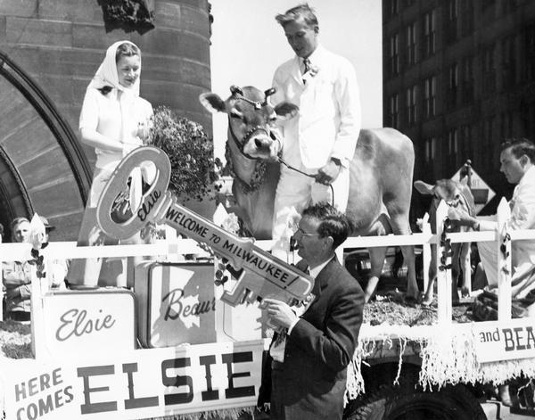 Alice in Dairyland, Margaret McGuire, and Elsie the Cow and her calf Beauregard, stand on a parade float with two men in downtown Milwaukee, while a male official on the ground holds up a key inscribed: "Elsie - Welcome to Milwaukee!"