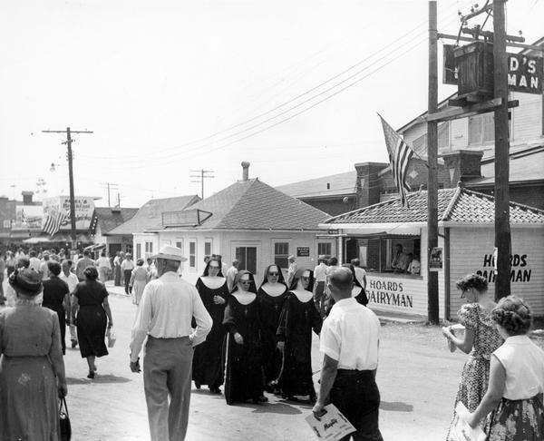 People saunter along the midway at the Wisconsin State Fair. A group of four nuns appears in the foreground, passing the Hoard's Dairyman booth.