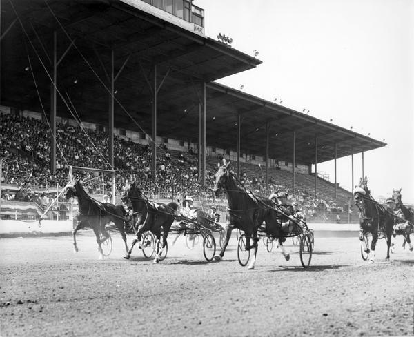 Harness racers are shown in action at the Wisconsin State Fair Park, with an audience watching from the grandstand.