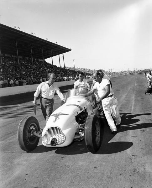 A race car is moved along the racetrack at the Wisconsin State Fair Park by its pit crew as a grandstand full of spectators watch.