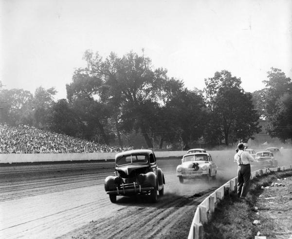 A photographer stands close to the Milwaukee Mile racetrack with camera poised to capture the action as the stock cars competing in the race speed by him. Spectators watch from bleachers on the other side of the track.