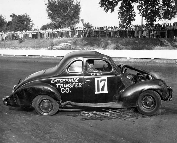A 1930's coupe sponsored by the Enterprise Transfer Co. stalls out on the racetrack after being disabled by an apparent collision. A crowd of onlookers stands behind a fence watching.
