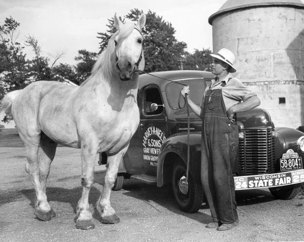 Young farmer Harvey A. Nelson, holding onto a large horse's lead, is standing next to an International truck at Gray View Farm. The truck's front bumper bears a bumper sticker promoting the Wisconsin State Fair, and a plate attached to the top of the license plate mentions the Racine County Fair.