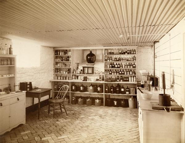 Crocks and bottles of various sizes line shelves, while beakers and mortars and pestles rest on counter tops in the brick-floored pharmaceutical lab at the Wisconsin State Hospital for the Insane (Mendota Mental Health Institute).