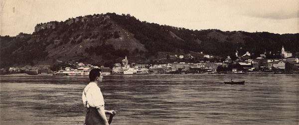 View across the Mississippi River of Eagle Bluff and Fountain City, with a man in the foreground. The steamer "U.S. Gen. Barnard" is at a landing and a rowboat is in the river.