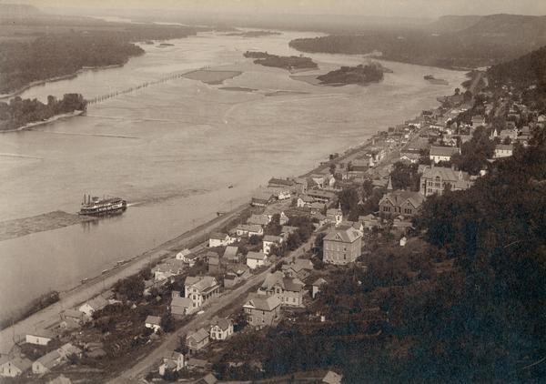 Elevated view of the city and the Mississippi River from the bluffs above the city. Photograph was taken after 1902 when the Buffalo County Training School building was erected. A steamboat is on the river.
