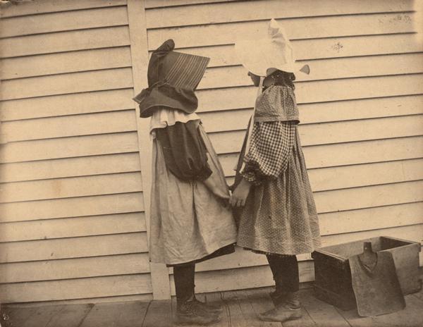 Two young girls in bonnets standing on a porch facing each other and holding hands. These may be daughters of Gerhard Gesell, the photographer, he often posed them in pictures to document their clothes and playtime.