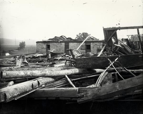 Boy standing among the wreckage of various farm structures and implements apparently after a wind storm or tornado.