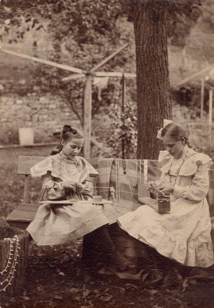 Two girls seated on a bench outdoors and working inkle looms.