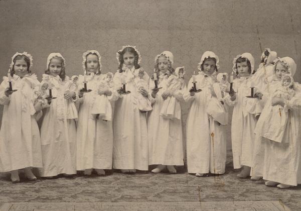 Group of little girls posed wearing nightgowns. Each holds an identically dressed doll and a candle.