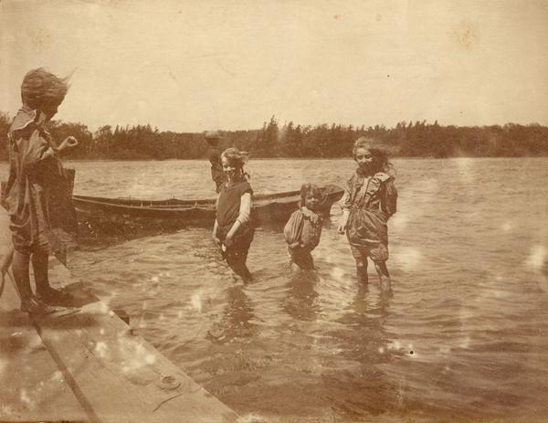 Three girls wade in a lake near Alma while another watches from a pier. A boy fishes from from a boat nearby.