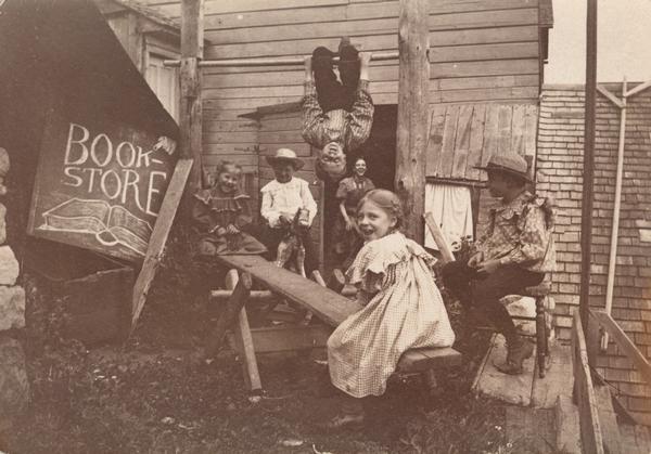Five children play in the yard while a young woman watches from the doorway. A boy hangs from a cross bar; a boy rides a rocking horse; two girls sit on a teeter totter; a hand-lettered sign at the left of the image reads "Book Store."