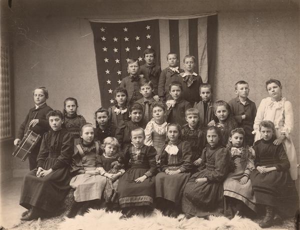 Group of twenty-five children posed in front of a 38-star United States flag. One girl holds an African American doll and one boy holds a drum. Note: The 38-star flag was valid from 1877 to 1890.