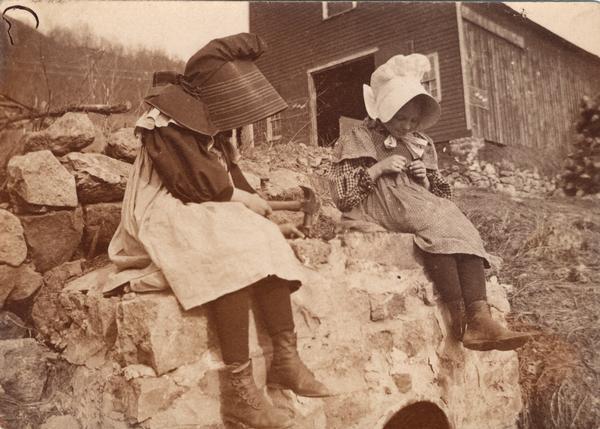 Two bonnetted farm girls sit on a stone wall, apparently cracking nuts.