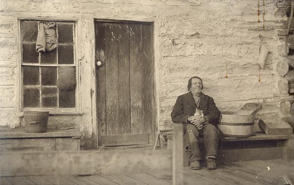 Abraham Schmocker, a "hermit" who lived in "Stumptown" near Alma, seated on a bench outside his log cabin, holding a kitty.