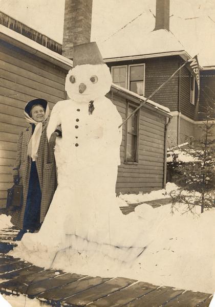 Winter scene with Wilma Gesell, daughter of Gerhard Gesell, stands on a board sidewalk arm-in-arm with a snowman. The snowman holds a Wisconsin pennant.