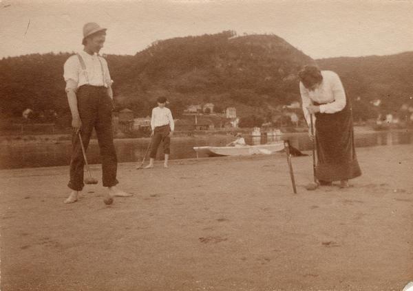 Three people playing croquet on the banks of the Mississippi River near Alma, Wis. A woman is sitting in a rowboat with a dog nearby in the background.