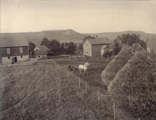 Elevated view across field with large haystacks towards a woman sitting in horse-drawn carriage in a farmyard.