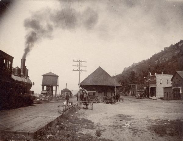 CB&N Railroad locomotive entering town. Railroad station in center.