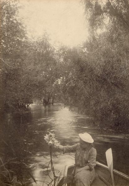 One of Gerhard Gesell's daughters (Wilma or Bertha) sitting in a rowboat holding a bouquet of Mississippi River water lilies. She is seated in the stern, twisting to the left, with that arm upraised holding the flowers out over the water. There is a raised oar in the foreground, with a tree-lined river in the background.