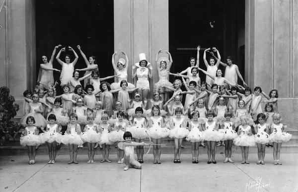 A large group of female dancers of varying ages pose in a variety of costumes at the Kehl School of Dance. The young girls in the front row wear dresses adorned with numbers, and the girl kneeling in front wields a bow and arrow.