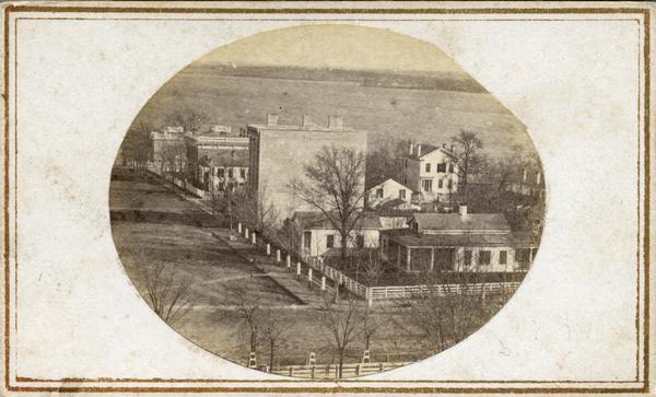 South Wisconsin Avenue and West Main Street. Lake Monona is in the background and the Lucius Fairchild and David Atwood houses are at the upper left. The residence of Simeon Mills faces the Capitol Square. The name of South Wisconsin Avenue was changed on December 1, 1877, to Monona Avenue; it's now Martin Luther King Jr. Blvd.