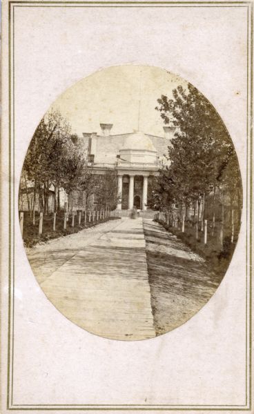 Wisconsin Territorial Capitol (first in Madison) viewed from State Street. The back wall of the east wing of the replacement capitol (second on the site) can be seen behind the Territorial Capitol. A smaller building, possibly a construction shack, is to the left.