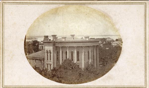 East (King Street) wing of the third Wisconsin State Capitol and the south wall of the Territorial Capitol (at the left) taken from the roof of the building at the corner of West Main and South Carroll Streets.  The east wing was the first phase of that building constructed.  Lake Mendota is in the background. The top story and clock tower of City Hall are visible just above the Capitol. Residences at the corner of East Gilman and North Pinckney Streets are in the upper right.