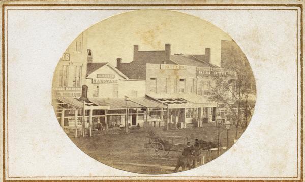Buildings located in the first block of East Main Street. Businesses in the image include Gleason Hardware and the Madison Institute. Two men on horseback and one man standing are in the forEground. Fuller probably made this image from his studio or the porch roof of the U.S. Hotel.