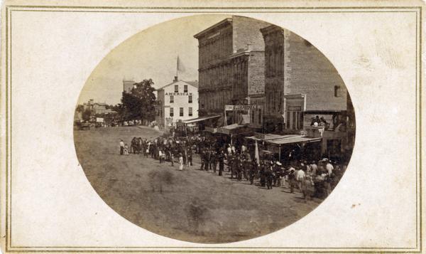 Elevated view of people gathered on Pinckney Street, possibly waiting for a parade. In the distance another group of people, some of whom are carrying a banner, are approaching. Other bystanders are seated on rooftops. A large flag is flying on the roof of the American Hotel.