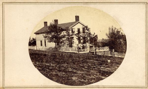 Exterior of a residence, probably of 1 East Gilman Street. The yard is enclosed by a fence. A dog is standing in front of the house.