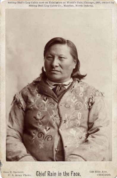 Waist-up studio portrait of Chief Rain in the Face, member of the Sioux tribe. He is wearing an elaborately tooled leather or suede coat.