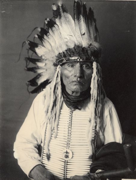 Studio portrait of Candi Tanka (Big Tobacco) with headdress and breastplate, holding pipe. Part of Siouan (Sioux) and Yankton Tribes