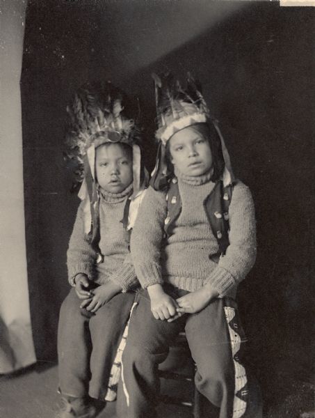 Studio portrait of two boys, Wilbert and Rosses Deon, both in partial native dress with headdresses. Part of Siouan (Sioux)and Brule Tribes