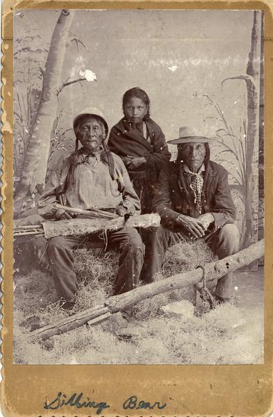 Studio portrait in front of a painted backdrop of three Native Americans, two older men and a young girl, possibly Winnebagos. "Silbing Bear" is written on the bottom of the photograph.