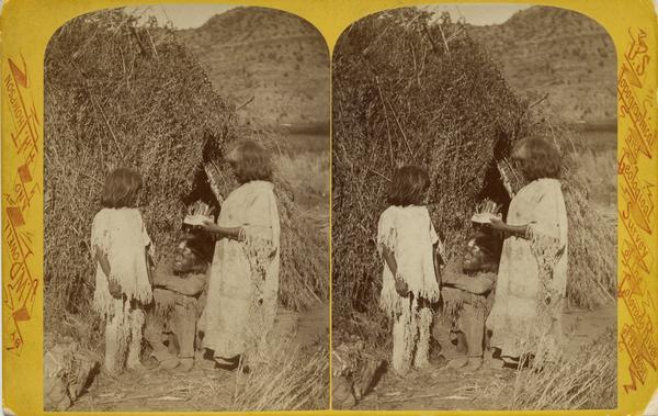 Stereograph image of a Native American man and woman with a young child, members of the Ute Indian Tribe, standing in front of a grass structure.