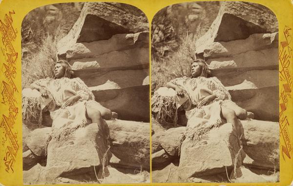 Native American woman, a member of the Ute Indian Tribe, seated on a rock outcropping in traditional costume.