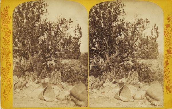 Stereograph of Native Americans, members of the Ute Indian Tribe, seated around a fire with a stick tripod structure holding a metal cooking pot.